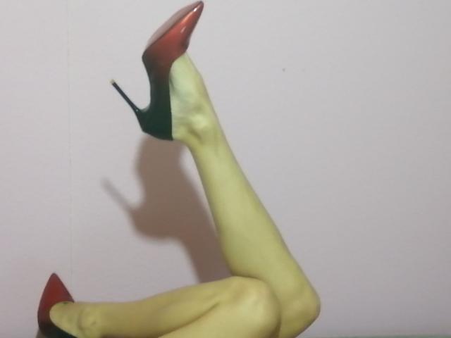 LaFrancaise's yellow-green leg is raised, wearing red-to-black high heels with green soles, shadow on pale wall. Boobs and pussy aren't visible, just the leg and shoes.