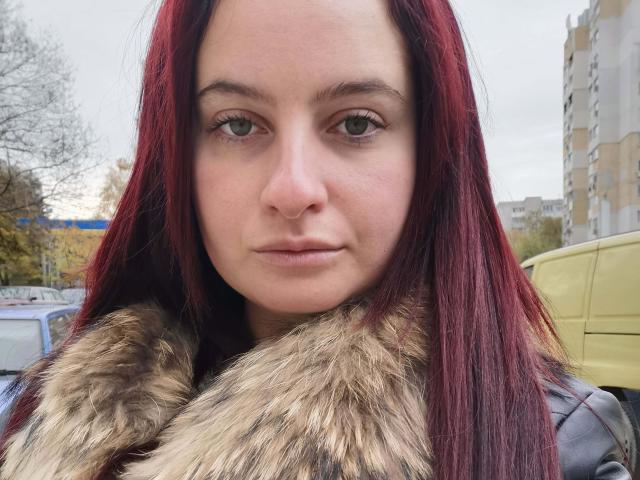 MarielaHot69 has red hair, green eyes, and a serious face. She wears a fur-lined jacket. Her nose is small and her skin is fair. Her tits are not visible.