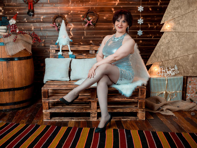 AlbertaAllen, middle-aged woman with curly brown hair, wearing a sparkly blue dress, black flats, and sheer stockings, sitting on a wooden bench with a fluffy white pillow.