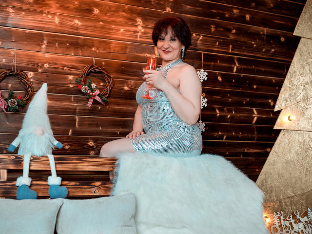 Alberta Allen, vestindo um vestido azul brilhante, sentada em um sofá branco coberto de pele, segurando uma taça de champanhe, sorrindo, com pequenos gnomos e uma parede de madeira ao fundo..