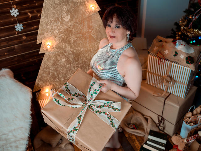 AlbertaAllen with short curly brown hair, wearing a shiny silver dress, holding a big brown gift with a green ribbon, standing by a decorated Christmas tree and presents.