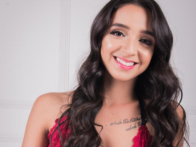 TifanyAngel mit langen, welligen, dunkelbraunen Haaren, lächelnd mit pinkem Lippenstift, trägt einen roten Spitzen-BH und zeigt ein Tattoo auf ihrer Brust. Sie ist ein süßes, sexy Mädchen..