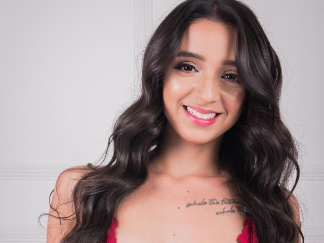 TifanyAngel has long, wavy dark brown hair, a cute smile showing pink lipstick, and a tattoo on her chest. She's wearing a red lace bra.