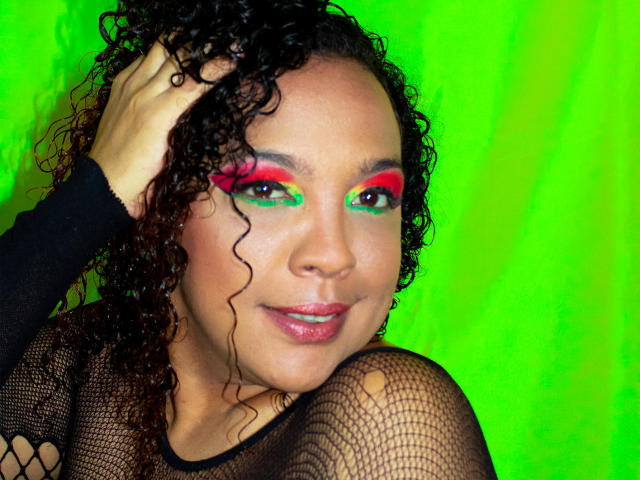 DevoraXPenelope with curly black hair, wearing fishnet top, has bright green, red, and pink eye makeup, smiling with shiny pink lips, hand in curly hair, against neon green background.