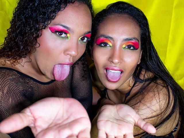 DevoraXPenelope and another DevoraXPenelope with curly and straight dark hair, sticking out their tongues, wearing colorful eye makeup and black mesh tops, showing their hands in the foreground.