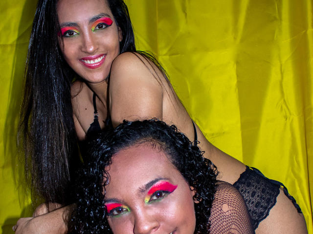 DevoraXPenelope, com longos cabelos pretos e sombra vermelha, sorrindo, vestindo lingerie de renda preta, inclinada sobre DevoraXPenelope, que tem uma maquiagem semelhante, com fundo amarelo, ambas mostrando os seios..