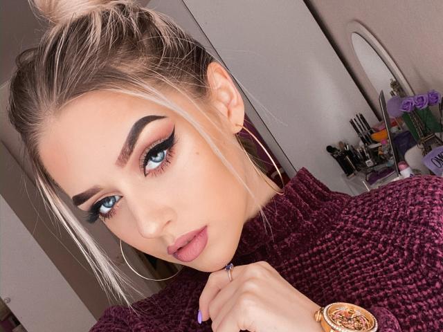 MaryVi met blond haar in een knot, blauwe ogen, dikke zwarte wenkbrauwen en roze lippen, draagt een paars gebreid top, gouden oorringen en een gouden horloge, en kijkt naar de camera..