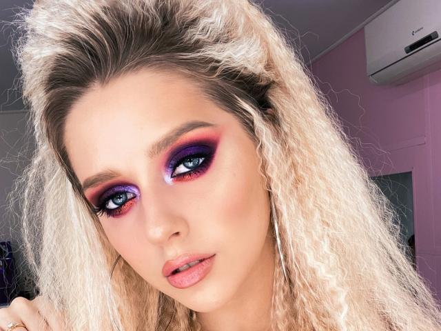 MaryVi mit blonden, lockigen Haaren, lila und rosa Lidschatten, blauen Augen, rosa Lippenstift und leicht geöffnetem Mund, die in die Kamera schaut. Klimaanlage im Hintergrund..