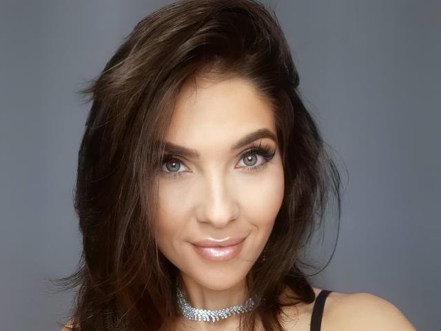 LovelyKinsley has dark brown, wavy hair, blue eyes, and a shiny pinkish-nude lip. She's wearing a silver choker and black top. Her tits are not visible.