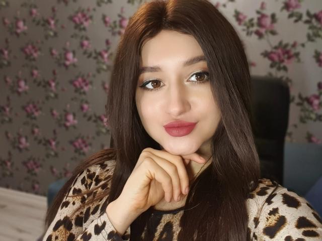 LailaNoire, con lunghi capelli castani, occhi marroni e rossetto rosa, indossa una maglietta a stampa leopardo, appoggiando il mento sulla mano e sorridendo dolcemente..
