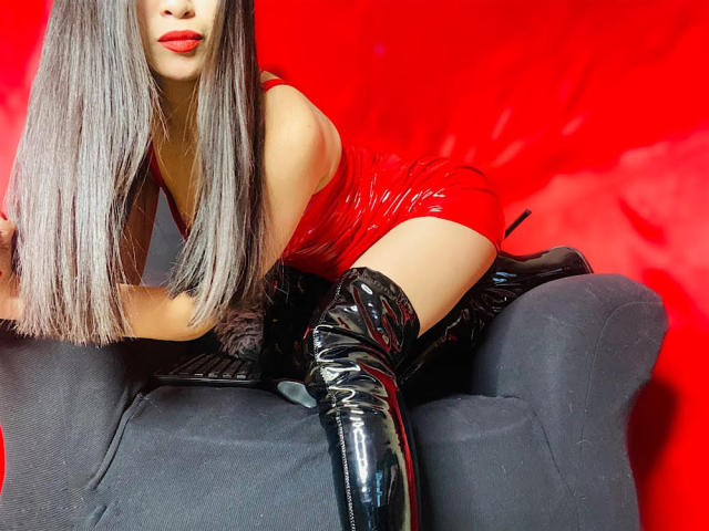 MistressCruelX com cabelos grisalhos longos, vestido de látex vermelho e botas pretas que vão até a coxa, ajoelhada em uma cadeira preta, exibindo seu traseiro e lábios vermelhos, contra um fundo vermelho..