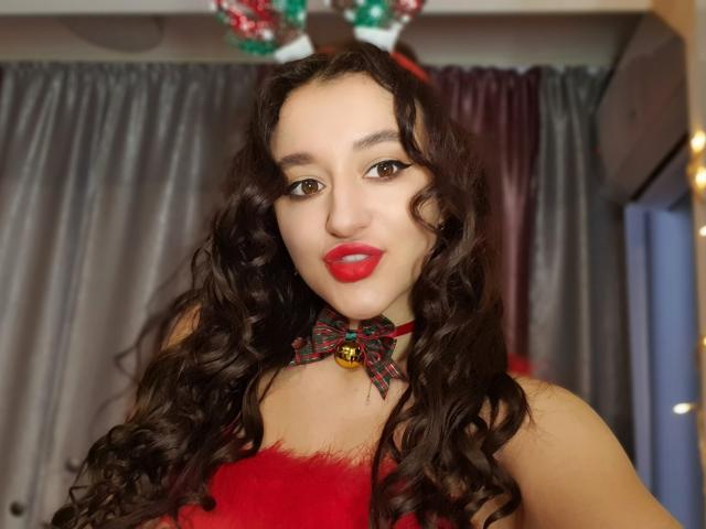 LailaNoire met lang, krullend donker haar, rode lippenstift en een rode, pluizige top, draagt een rode en groene lintenketting met een belletje, en kijkt met een lichte glimlach naar de camera..