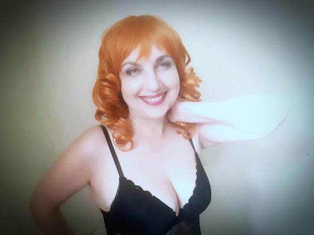 Stellacool with orange curly hair, smiling, wearing a black lace bra, showcasing her tits, pale skin, hand on neck, light background, and bright smile.