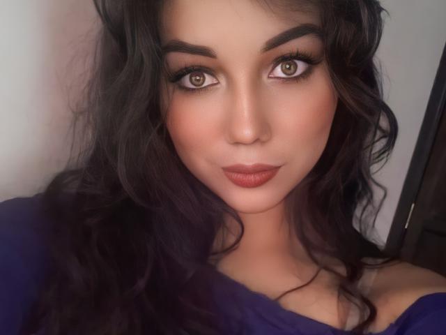 SquirtingCouple has dark, wavy hair, brown eyes, and full, pink lips. She's wearing a purple top, showing off her cute, sexy face with a slight, confident smile.