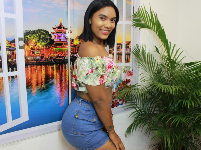 Monserrat Oliveira, di carnagione scura, ha un sorriso grazioso, indossa una blusa a fiori con una spalla scoperta e pantaloncini di jeans blu, ed è in piedi vicino a una finestra con il riflesso colorato di un tempio asiatico. Una pianta verde si trova a destra..