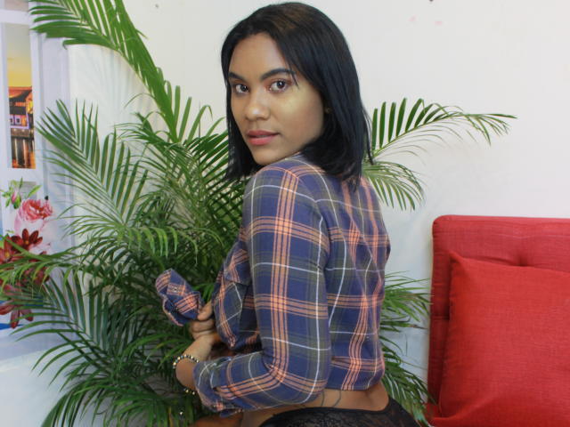 Monserrat Oliveira, mit hellbrauner Haut und schwarzem, schulterlangem Haar, trägt ein blau-kariertes Hemd, schwarze Spitzenunterwäsche und blickt über ihre Schulter, während sie neben einem roten Sofa steht..