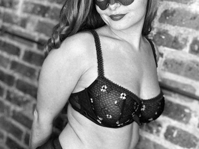 Reggiseno nero con pizzo floreale, che copre i seni di LustFrenchX, davanti a un muro di mattoni. Indossa una maschera, ha i capelli ondulati e un sorriso sicuro. Foto in scala di grigi..