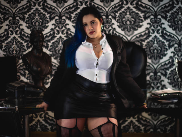 LissaClaire, com cabelo azul, jaqueta preta, camisa branca, saia de couro preta, meias de renda e seios grandes, sentada em um escritório escuro, olhando para a câmera..