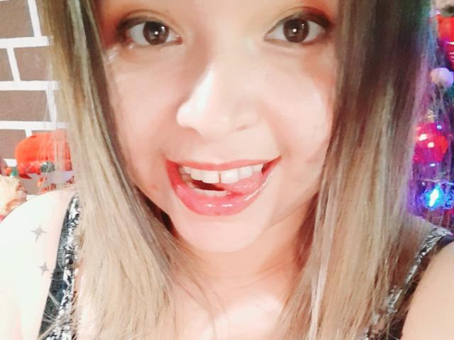 Myalewwis has a shiny red-lipped smile, showing her white teeth, with her tongue slightly out. She has brown eyes, blonde hair, and is wearing a black patterned top.