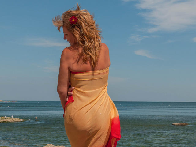 AdamaXbella with blonde wavy hair, red flower in hair, yellow and red draped fabric covering her tits and ass, standing by the ocean, blue sky, and calm water.