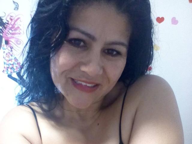 Un selfie de SweetieOnline con cabello negro ondulado, ojos marrones y una sonrisa encantadora, mostrando los hombros desnudos y una correa negra, con lápiz labial rojo y un discreto lunar en la nariz..