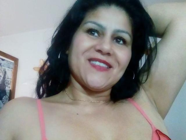 SweetieOnline has medium brown skin, black wavy hair, and is smiling with red lipstick. She's wearing a pink bra, has small tits, and is raising her arm.