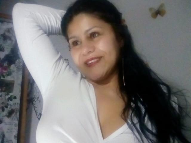SweetieOnline con lunghi capelli neri, una maglietta bianca a V, un braccio alzato, che sorride, mostrando il seno, pelle color nocciola, occhi marroni, piccoli orecchini a cerchio e un leggero rossetto rosa..