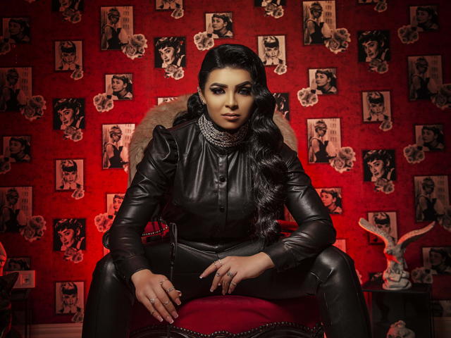 Devasha with dark wavy hair, wearing a black leather outfit, sitting on a red chair, looking serious, surrounded by black-and-white photos of a woman's face on red wallpaper.