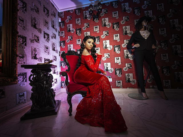 Devasha, vestindo um vestido vermelho com detalhes em renda e ombros descobertos, está sentada em uma cadeira escura e ornamentada, com as mãos no peito, e à sua direita, há um manequim vestido de preto, em um quarto elegante..