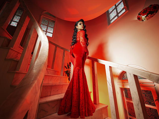 Devasha stands in a red-laced, form-fitting dress, showing off her ass and long black hair, on a marble staircase, with a red light casting a sexy glow.