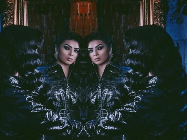 Three mirrored Devashas with dark, shiny hair, heavy makeup, and black puffed jackets, standing with arms crossed, reflecting each other's serious, seductive faces.