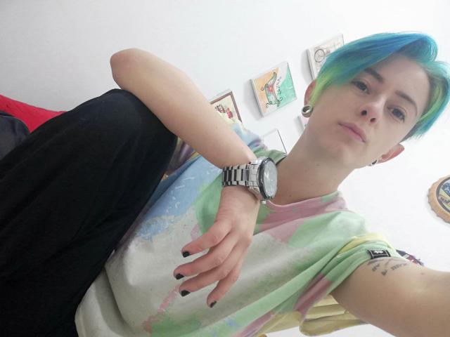 RedFury, con cabello corto azul verdoso, vistiendo una camiseta con estampado tie-dye y pantalones negros, tiene esmalte de uñas negro, un reloj de plata y aretes grandes, y está sentada con una expresión relajada..