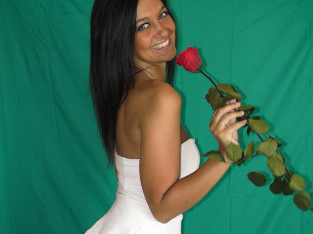 SelinaKylla, con i capelli neri, sorridente, che tiene una rosa rossa, indossa un asciugamano bianco senza spalline, mostrando la schiena e i glutei, sfondo verde, capelli lunghi e lisci, sorridente, che guarda oltre la spalla..