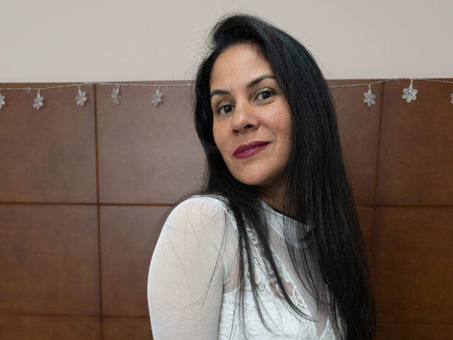 RoraimAnaggie heeft lang zwart haar, donkere ogen en roze lippenstift, en draagt een witte, doorschijnende top. Op de achtergrond is een bruine muur met tegels te zien, en zilveren decoraties..