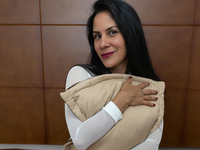 RoraimAnaggie, with long black hair, white shirt, and red lipstick, hugs a beige pillow, smiling at the camera, showing off her cute ass. Her nails are painted red.