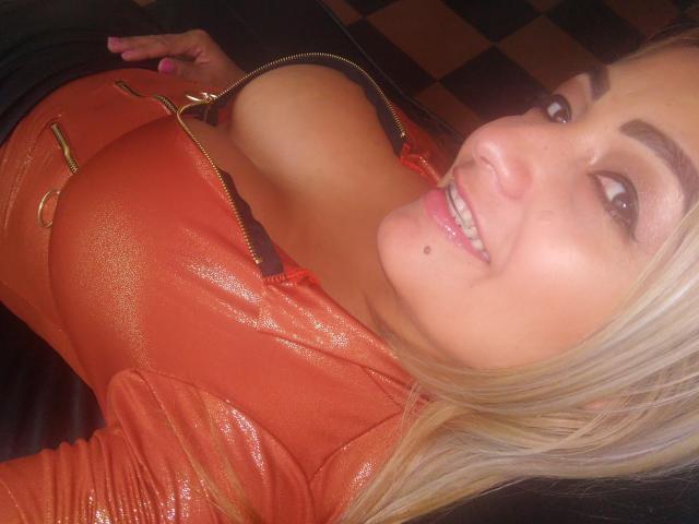 SexyBush with large tits in a shiny orange leather top, smiling at the camera, has blonde hair, a mole on her chin, and pink nail polish.