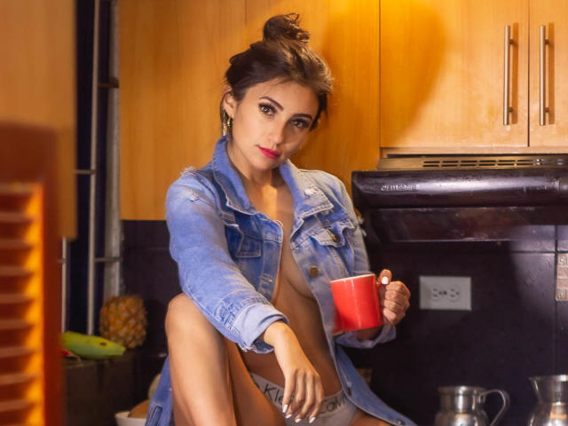 SammyHoffman with dark hair in a bun, wearing an open blue denim jacket, holding a red mug, sitting on a kitchen counter, showing her tits, with a pineapple in background.