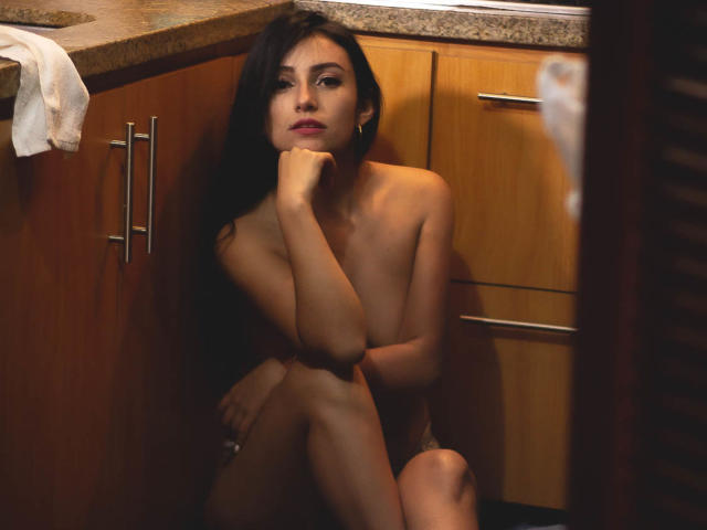 Nude SammyHoffman with long dark hair, sitting in a kitchen, arm over tits, hand on chin, legs crossed, wood cabinets, granite countertop, dim lighting, looking at camera.