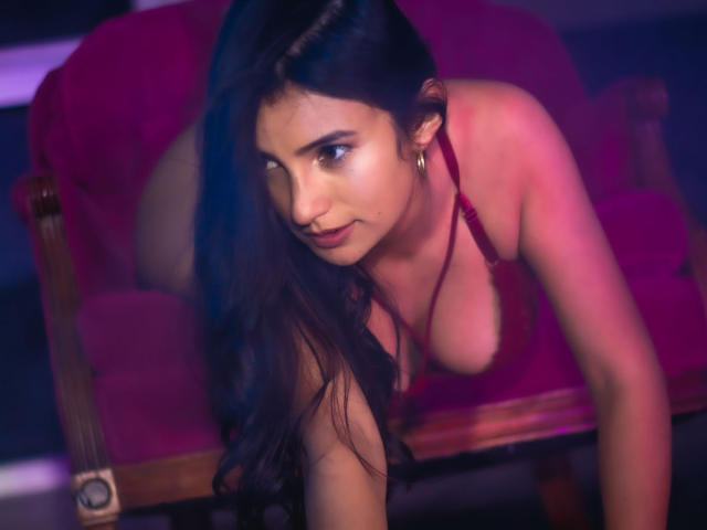 Sammy Hoffman, con cabello largo y oscuro, lencería roja y senos grandes visibles, inclinada hacia adelante sobre un sofá rojo, mirando seductoramente a la cámara, en una habitación iluminada con luz púrpura..