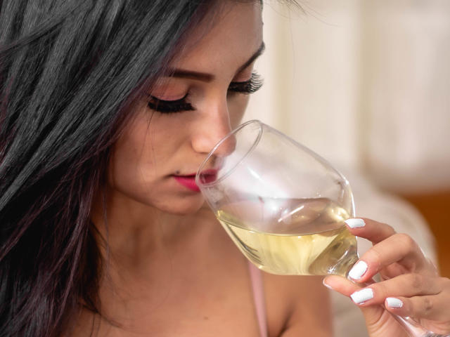 SammyHoffman with long black hair, pink lipstick, and white nails, sips from a glass of white wine, eyes closed, pink top, soft light, subtle makeup.