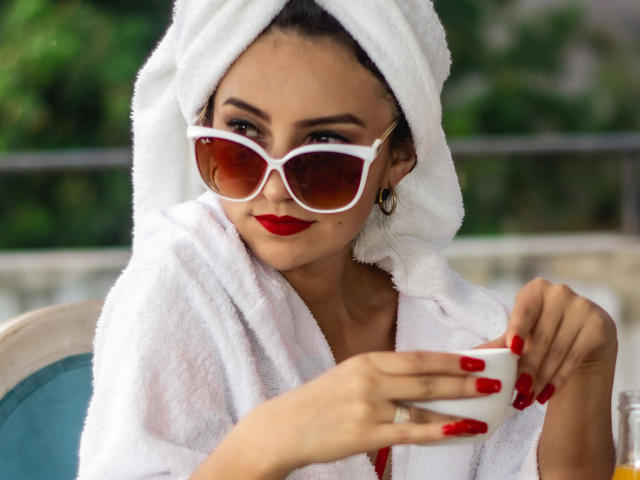 Sammy Hoffman, com uma toalha branca na cabeça, usando batom vermelho e óculos de sol brancos, segurando uma xícara branca com unhas vermelhas, parecendo adorável e relaxada..