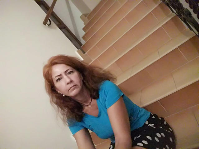MilayaDFlow with red hair, blue shirt, and polka-dot black pants, leaning on stairs, looking annoyed, tits slightly visible under shirt, medium build, fair skin, slight smile.