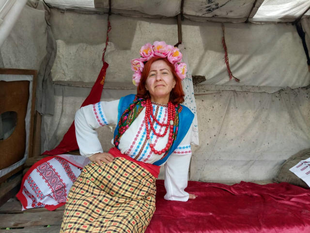 MilayaDFlow con capelli rossi, una corona di fiori rosa, una maglietta blu, maniche bianche, una collana di perline colorate, perline rosse, una gonna a quadri gialla, un tessuto bianco e rosso, seduta su un tessuto rosso..