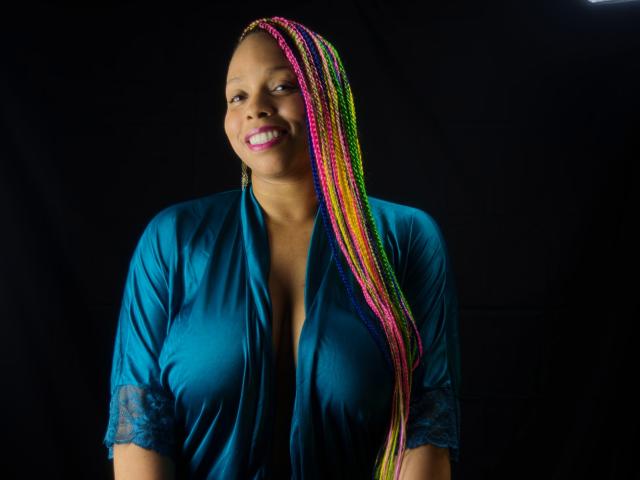 OpheliaWinter with colorful braids, smiling, wearing a teal, lace-trimmed, open-front top, showing her tits, against a black background.