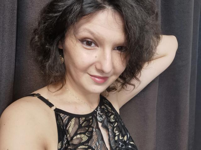 AmethystCharm with curly black hair, wearing a black lace top, has her arm behind her head, smiling with pink lips, brown eyes, and small nose, against a dark curtain.
