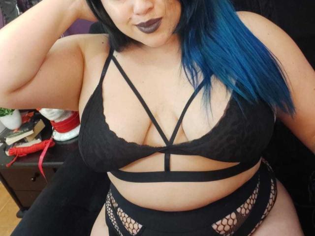 LissaClaire with blue hair, black lace bra, strappy top, and fishnet bottoms, showing her big tits and fat ass, with dark lipstick and a hand in her hair.