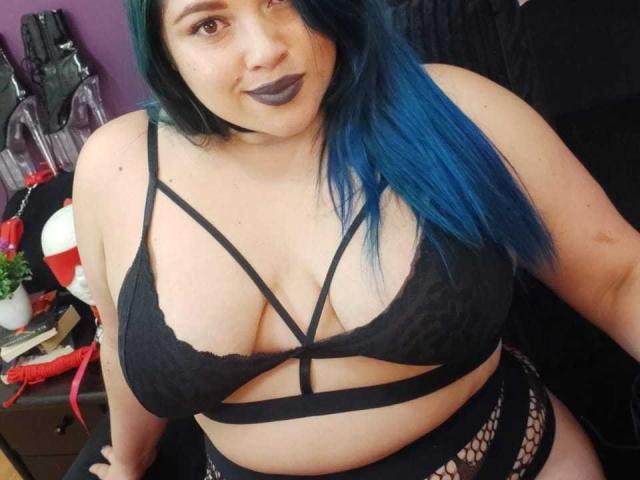 LissaClaire with blue hair, black lace bra showing her big tits, strappy design, and fishnet bottoms, sporting dark lipstick, sitting down.