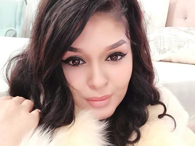 LexyRene tiene el cabello oscuro y ondulado, ojos marrones con delineador de ojos llamativo, y lleva un abrigo de piel blanco, con una sonrisa sutil, sosteniendo su cabello en el lado izquierdo..