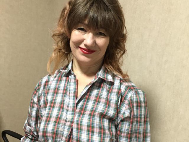 SonyaAir with wavy brown hair, wearing a red plaid shirt, smiling with red lipstick, her tits slightly visible under the shirt, standing against a beige wall.