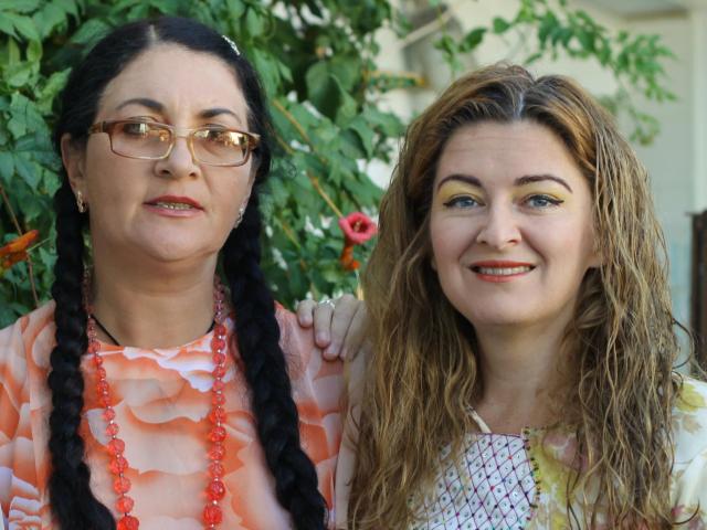 Dos mujeres, OzzyHelen con trenzas negras y gafas, vestida con un vestido floral rosa y un collar de cuentas rojas, está de pie junto a OzzyHelen con el cabello castaño mojado y sombra de ojos amarilla..